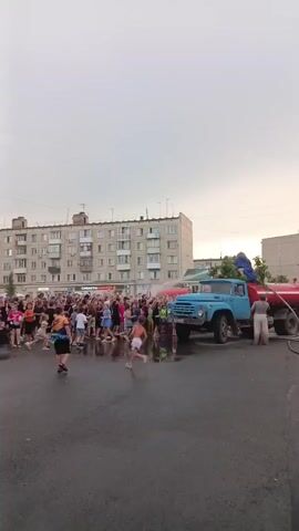 A crowd of people, many of them young, are gathered in an outdoor area in front of a multi-story apartment building. The ground is wet, suggesting recent rain or a water event. A blue truck, possibly a water truck, is parked on the right side of the frame. A person is standing on the back of the truck, and another person is standing next to it. Some people in the crowd appear to be interacting with the truck or the water. The sky is overcast.