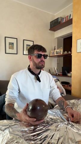 Here is a concise description of the video:
A man is sitting on a couch in a living room with walls painted light peach. The man is wearing a white long-sleeve shirt, sunglasses, a black bandana around his neck, a silver bracelet, and tattoos on his arms. He is sitting at a table that is covered in aluminum foil. He is holding a large, round, chocolate easter egg. He drops the chocolate egg onto the table and it breaks in half. He puts his index finger and thumb together in an Italian hand gesture. There are two framed pictures on the wall behind the couch and a bookshelf in the background. A person's legs are visible to the left of the man.