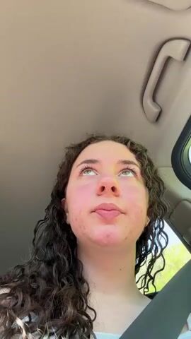 A young woman with curly brown hair is in a car. She is wearing a seatbelt and is looking up, then looks into the camera. There is music playing. The interior of the car is visible, including the ceiling handle and a window with sunlight coming through. The woman has light skin with some blemishes.