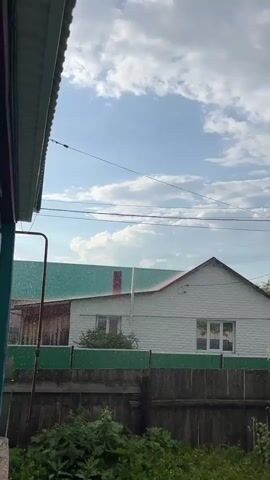 A cloudy sky with scattered white clouds is visible above a residential area. In the foreground, a portion of a house's overhang is seen on the left. A wooden fence runs horizontally across the middle ground, with lush green foliage in front of it. Behind the fence, two houses are visible. The house on the left has a green roof, and the house on the right has a white exterior and a red-trimmed roof. Rain is falling, visible as streaks against the sky and houses. Power lines are strung across the sky.