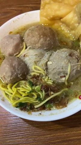 A close-up shot captures a bowl of bakso, a popular Indonesian meatball soup, presented on a wooden surface. The bakso, featuring large, spherical meatballs, is submerged in a clear, savory broth. Thin, white rice noodles and bright yellow egg noodles are intertwined within the soup, offering a textural contrast. Crispy fried wontons, golden in color, are arranged on the side of the bowl. Garnishes of chopped green onions and fried shallots add visual appeal and aromatic depth to the dish. A bright yellow spoon, laden with a dollop of red chili sauce, hovers above the bowl, suggesting the act of seasoning the soup. The lighting is warm and inviting, hinting at a casual mealtime. The overall impression is one of a delicious and satisfying culinary experience, likely enjoyed in a restaurant or home setting in South Tangerang, Indonesia. No visible text is present.