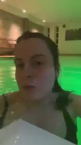 A woman is in a swimming pool with green-tinted water, wearing a black swimsuit and holding a white towel. The room is decorated with stone walls and has artificial lighting, giving the water a green glow. The woman looks directly at the camera with a neutral expression.