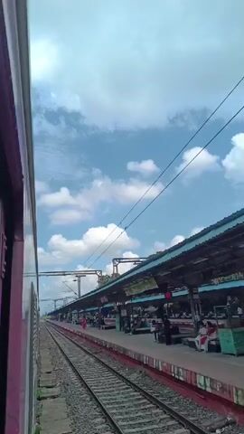 A train station platform is visible from inside a moving train. The platform is covered by a long, arched roof supported by columns. Several people are sitting on benches under the roof. Train tracks run alongside the platform. The sky is blue with scattered white clouds. The train car to the left is painted white and maroon.