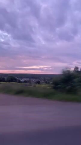 The video shows a blurred, passing view of a rural landscape. The sky is cloudy with hues of purple and pink, suggesting either sunrise or sunset. In the distance, there are hints of buildings and trees. The foreground is dominated by the blur of green vegetation and a road with a white guardrail. The camera appears to be moving, possibly from a vehicle, indicated by the motion blur. There are no people or distinct objects visible with enough clarity to describe. The overall impression is of a quick drive through the countryside at dusk or dawn.
