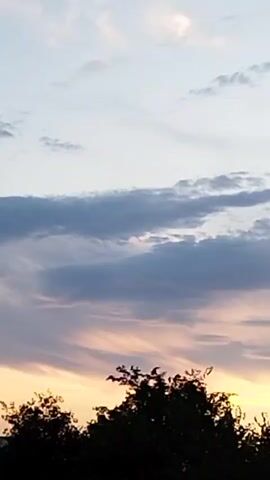 The video shows a time-lapse of a sunset. The sky is filled with clouds, which are illuminated by the setting sun in shades of orange and yellow. Silhouettes of trees are visible at the bottom of the frame.