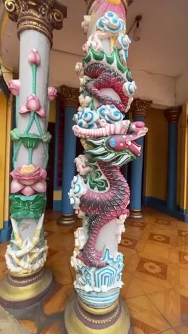 The camera pans across the entrance to a Buddhist temple or religious site.  Two decorative columns are visible, one sculpted with a dragon and the other with lotus flowers. The dragon is predominantly red and green with white clouds, and the lotus flowers are pink and green.  The entrance is framed by blue pillars with gold trim and dark wood double doors.  A golden framed painting hangs above the entrance. A statue of a deity holding a long blade stands in a niche to the right. The floor is tiled in a diamond pattern of brown and yellow.