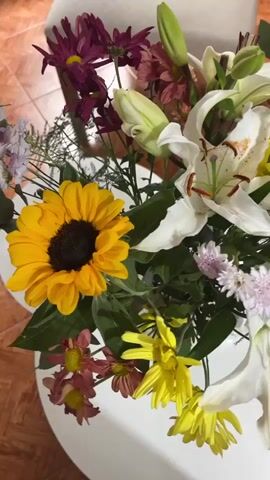 The video shows a floral arrangement in a white round vase. The flowers include a sunflower, white lilies with brown stamens, yellow chrysanthemums, small purple aster-like flowers, and some red-brown chrysanthemums. Some of the lilies are still buds. The vase sits on a table with a red-tiled floor. Later in the video, the table is situated on a wooden floor near a white chair and sofa. The camera circles the arrangement, providing a 360 degree view.