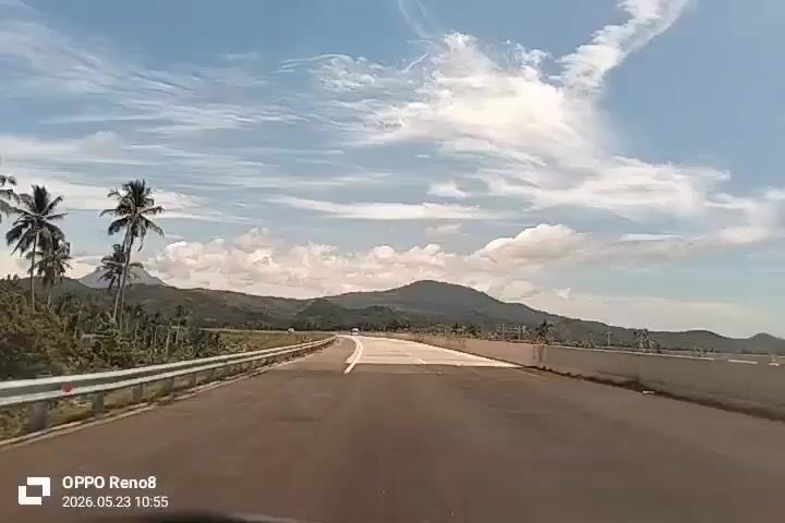 The video shows a vehicle's point of view driving on a long, straight highway. The road is paved and has a white line marking the lanes. On either side of the road, there are guardrails. Beyond the guardrails, there are trees and mountains in the distance. The sky is blue with scattered white clouds. The video is taken with an OPPO Reno8 phone, as indicated by the watermark at the bottom of the frame. The timestamp on the watermark indicates May 23, 2026, at 10:55 AM.