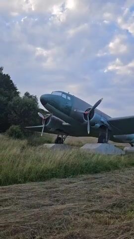 A vintage, dark green airplane with twin propellers is parked on a concrete base in a grassy field under a cloudy sky. The camera pans across the aircraft's fuselage and wing. A person with blonde hair, wearing a black t-shirt and blue jeans, walks away from the camera towards the airplane. The airplane has a red star emblem on its tail fin. The surrounding environment consists of tall grass and trees in the background.