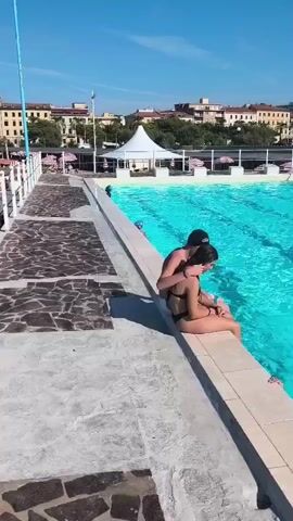 A couple sits on the edge of a swimming pool. The man has his arm around the woman. The woman is wearing a black bikini and the man is wearing a dark baseball cap and dark shorts. They are facing the water, and the woman's feet are in the water. The pool is large and bright blue. There are other people in the pool, some swimming and one on the edge of the pool. In the background, there are buildings and trees under a clear blue sky. The poolside area has lounge chairs and umbrellas.