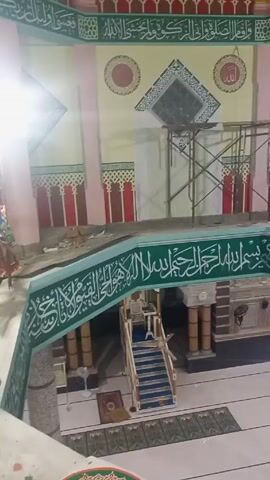 The video shows the interior of a building, possibly a mosque, under construction or renovation. The camera pans upward, revealing the multi-tiered architecture, including ornate Islamic calligraphy and geometric patterns painted on the walls. There is scaffolding in place, suggesting ongoing work. The camera then focuses on the ceiling, which features a dome with decorative elements. The color scheme is predominantly light, with accents of green, red, and gold.