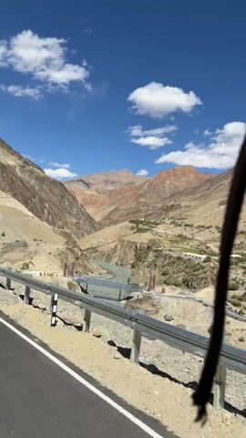 The video begins with a wide shot of a scenic mountain valley with a river flowing through it. The mountains are barren and dusty, with some sparse vegetation visible. A bridge is under construction across the river, with cranes and construction equipment present. The camera then zooms in on the construction site, showing more details of the bridge's progress. The shot then shifts to inside a vehicle, showing the legs and feet of a person. The person is wearing hiking boots and dark pants. The vehicle appears to be moving, as evidenced by the slight blur in the background. The video ends with a close-up of the person's legs and feet.