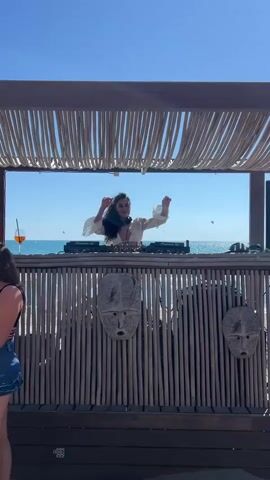 A woman is DJing at a beach party. She is wearing a white top and has long, dark hair. She is standing behind a DJ booth made of wood and decorated with tribal masks. The ocean is visible behind her, and the sky is blue. There is a drink with an orange garnish on the counter to the left of the DJ booth. The woman is raising her hands in the air, and appears to be dancing to the music. The video is shot from a low angle.