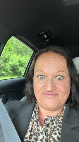 The video features a woman in the driver's seat of a car, looking directly at the camera. She has dark hair, green eyes, and is wearing a leopard print top under a dark jacket, with a seatbelt across her chest. A hint of a smile is on her face. Through the rear window, green trees and a white road sign are visible in the distance. The camera then pans left, showing the car's interior, including the seatbelt and the view outside the passenger window, which displays blurred green trees and a road, indicating the car is moving. The pan continues to the front passenger side, where a side mirror and an "AIRBAG" label on the car's pillar are visible, with more green foliage and the road passing by outside. The scene takes place during the day.