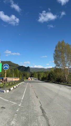 The video captures a view of a road leading into the town of Abaza, Russia, under a bright blue sky with scattered white clouds.  The camera pans upward to showcase the town's sign, which includes the name "АБАЗА" in Cyrillic letters, the founding year 1856, and an emblem featuring a silhouetted figure holding a spear. The Russian, Khakassian, and Abazin flags are waving atop the sign. The landscape includes lush green trees, mountains in the background, a partly visible moon, and road signs indicating lane directions.