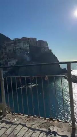 The video shows a picturesque scene of a coastal village nestled on a hillside, overlooking the sea. A woman in a patterned top and black pants stands by a metal railing, looking out at the view. Beside her is a phone attached to a tripod, presumably used for filming. Another woman, wearing a light brown jacket and sunglasses, enters the frame and walks to the left. The setting is a stone walkway, and the weather appears sunny with a clear blue sky.