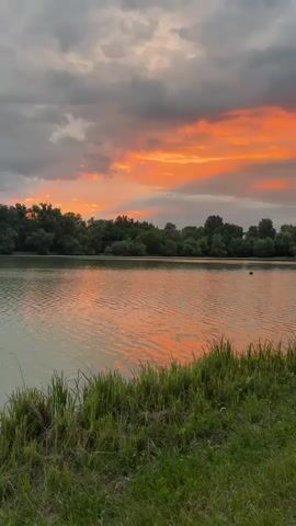 A still shot of a lake with trees in the background. The sky is filled with clouds, some of which are orange and red, suggesting a sunset. There is some grass in the foreground.