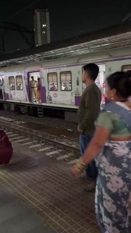The video shows a train station platform at night. A train with purple accents is stopped alongside the platform. People are on the train and some are standing in the doorways. Three people are walking on the platform. A woman in a blue and gray sari walks towards the left of the frame. A woman in an orange outfit with a blue backpack is also walking towards the left. A man in jeans and a green shirt is walking towards the right. The platform has red and gray tiles. A tall building is visible in the background.