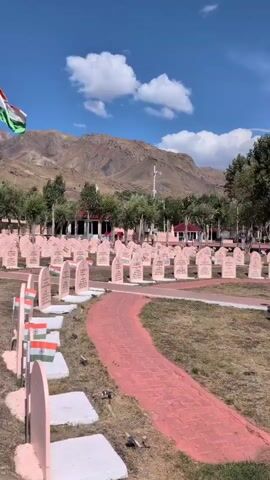 The video opens with a low-angle, panning shot of a cemetery. Rows of light pink tombstones, each with an inscription and a small Indian flag placed in front of it, fill the frame. A bright pink pathway curves through the cemetery. In the background, a large Indian flag flies from a tall flagpole under a clear blue sky with scattered white clouds. Beyond the cemetery, a range of rugged, barren mountains looms, and a few trees are visible. The overall impression is one of solemnity and remembrance.