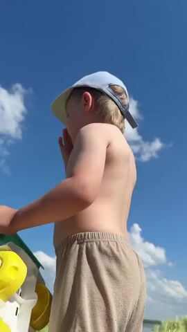 The video displays a low-angle shot of a young child wearing a light blue baseball cap and beige shorts, with their back to the camera. They are holding a colorful toy shovel with a yellow bucket attached. The child appears to be on a beach or similar outdoor location, evidenced by the clear blue sky with scattered white clouds and hints of greenery in the background, suggesting a sunny day. The child's skin is tanned, and their posture indicates they might be engaged in play, possibly building a sandcastle or digging. The camera angle focuses on the child from behind, emphasizing their silhouette against the bright sky. The overall atmosphere is one of childhood innocence and outdoor enjoyment, likely during daytime given the lighting. There is faint text visible on the toy, appearing to be in a Cyrillic script, possibly a brand name or warning label, but it is illegible. The provided location context "Новое Ильинское, Russia" suggests the setting is in Russia.