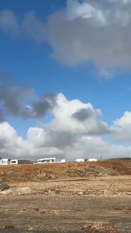 The camera is pointed towards several modern, white buildings perched on a small hill overlooking a flat, arid terrain. The ground is primarily brown and tan, but dotted with patches of red vegetation. A narrow, paved road lies in the foreground. The sky is a mix of clear blue and dynamic cloud formations, with some clouds appearing heavy and grey. A corner of a road sign is visible at the edge of the frame. The camera is likely moving slowly. The overall setting suggests a sparse, coastal landscape.