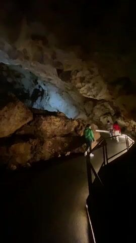 People are walking along a path inside a cavern. The path has a rail on one side and is illuminated by lights. The cavern ceiling is rocky. The video is somewhat dark.