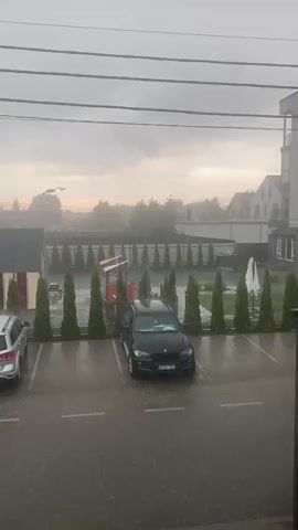 The video shows a parking lot outside an apartment building during a heavy downpour. The sky is overcast and the lighting is dim, suggesting it is either early morning or late evening. Rain is visibly falling, hitting the pavement and the roof of a covered parking structure. Two cars are parked in the lot, a silver SUV on the left and a black SUV under the covered structure on the right. A black sedan is partially visible in the foreground on the right. The apartment building is modern in design, with multiple stories and large windows. A row of conical evergreen trees lines the edge of the parking lot. The overall atmosphere is one of a severe weather event.