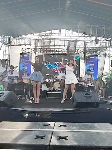Two women are on stage, performing with a band. The woman on the left wears a black top and a short, light-blue skirt, while the woman on the right wears a short, white dress and holds a microphone. Both are wearing high heels. A large screen is behind them, displaying a graphic logo with what appears to be the name of the show. The band is visible in the background, with musicians playing guitars and drums. The stage is set up under a covered structure, likely an outdoor venue. Several large speakers flank the front of the stage. The camera angle is from below, looking up at the stage.