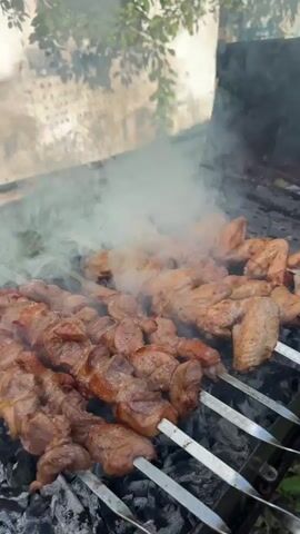 A close-up, eye-level shot shows skewers of meat cooking over a charcoal grill. Smoke billows from the grill, obscuring some of the food. In the foreground, several skewers are filled with cubes of reddish-brown meat, some of which are slightly charred. Behind these, another row of skewers holds what appear to be chicken wings, also cooking. The grill grate is visible, with glowing charcoal underneath. In the background, out-of-focus foliage and a light-colored wall suggest an outdoor setting. The lighting is natural, with the sun casting soft light on the scene. The overall impression is one of outdoor grilling.