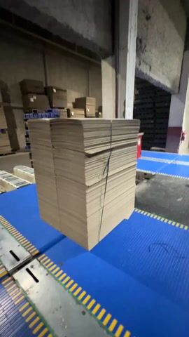 In an industrial setting, a large stack of flat, rectangular cardboard pieces, bundled together with black straps, sits on a blue conveyor belt with yellow striped edges. The camera slowly zooms in to reveal the layers of cardboard. Additional stacks of cardboard boxes are visible in the background on pallets.  The warehouse has bare walls and is lit by overhead fluorescent lighting.