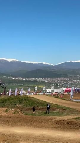 The video shows a motocross race on a sunny day with snow-capped mountains in the background. Several motocross riders are visible on a dirt track, jumping over a hill. There is a crowd of people watching the race, standing behind flags and banners. Two people are standing on the hill in the foreground, observing the race. The sky is clear and blue.