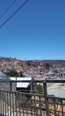 The video shows a townscape with houses built on a hill, filmed from a bridge or elevated walkway. The camera pans slightly, revealing a clear blue sky. The houses are closely packed, with corrugated iron roofs. In the distance, there is a line of hills or mountains. In the foreground, there is a metal railing, partially obstructing the view.