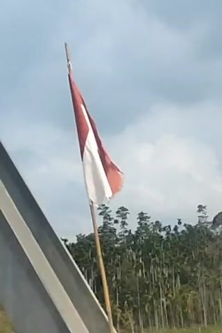 A red vehicle is driving on a road. The flag of Indonesia, red and white, is visible flying from a pole attached to the vehicle. Trees, some of which appear to be palm trees, are in the background. The sky is cloudy. The video also shows close-ups of the vehicle's exterior, including part of the grille and a word written on the side.