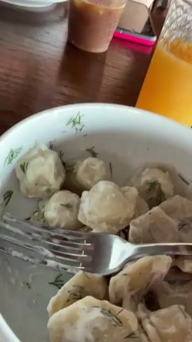 The video begins with a close-up shot of a bowl filled with dumplings in a creamy sauce, garnished with dill, and a fork resting on top. To the right of the bowl is a glass of orange juice. In the background, another drink in a small glass is visible. The camera then pans to the right, revealing a pair of eyeglasses on the table. The scene transitions to an outdoor setting, likely a restaurant or cafe patio, with a wooden floor and a covered roof supported by wooden beams. Several empty wooden lounge chairs are arranged on the patio. Beyond the railing, a body of water, possibly the sea, is visible, with inflatable water toys in the distance. The lighting suggests it is daytime.