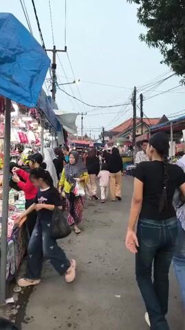 The video shows a busy outdoor market scene with many people walking along a narrow path. On the left side of the path, there are stalls displaying various goods, possibly clothing or accessories, under a blue tarp. Several people are browsing these stalls. On the right side of the path, there are more stalls and buildings in the background. The path is paved and appears to be an urban street. Overhead, there are numerous power lines and poles. The sky is overcast. The people in the video are dressed in casual clothing, and some women are wearing headscarves. The overall atmosphere suggests a lively marketplace.