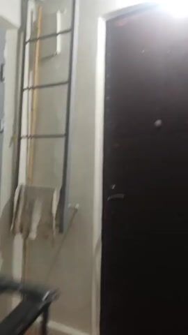 A video clip shows a light-colored wall with a dark brown door on the right. A tall, grey metal ladder leans against the wall on the left. A light-colored towel hangs from the lower section of the ladder. The camera pans upwards, revealing two swallows perched on the top rung of the ladder near the ceiling. A bird's nest is visible on a ceiling beam to the left of the left swallow. The camera remains focused on the swallows and the nest for the duration of the clip.