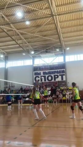The video captures a volleyball game taking place indoors. The scene is a gymnasium with a polished wooden floor, a high ceiling with exposed beams and fluorescent lighting, and large windows along one side. Spectators are visible in the background, seated in bleachers. The game involves two teams of young women, identifiable by their uniforms. One team wears green and yellow uniforms with the number 5 clearly visible on one player's jersey. The opposing team wears blue uniforms, with the number 15 visible on one player.

The activity depicted is a volleyball match, with players in various positions on the court, some actively engaged in play, others observing or preparing for the next move. The ball is visible in motion at different points in the short clip, indicating a rally in progress. The players exhibit athletic movements, including jumping and reaching.

Notable details include the setting being a sports facility, likely a school gymnasium, given the scale and amenities. The time of day is indeterminate due to the indoor lighting, but the overall atmosphere appears to be that of a competitive sporting event. There are no visible weather cues as the action is indoors. The emotions of the players are not explicitly discernible, but the intensity of the game is implied by their focused postures and actions.

Visible text in the background includes a large banner above the spectators that reads "МЫ ВЫБИРАЕМ СПОРТ" which translates to "WE CHOOSE SPORT". This reinforces the context of a sports event. The location context of Sharkan, Russia, further specifies the geographical setting of this sports activity.