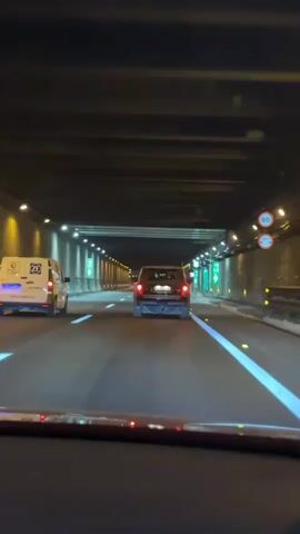 The video shows a view from inside a vehicle traveling through a well-lit tunnel at night. The road has multiple lanes, indicated by white lines. Ahead, there are two vehicles: a white van on the left and a dark-colored SUV in the center lane. Both vehicles are moving away from the camera. The tunnel walls are lined with lights that illuminate the passage. Green directional signs are visible on the right side of the tunnel. The interior of the vehicle from which the video is recorded is partially visible at the bottom of the frame, with red lights from the dashboard or other indicators reflecting on the windshield. The overall environment is dark except for the artificial lighting within the tunnel.