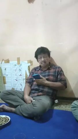 A man is sitting on the floor, leaning against a light-colored wall, and looking at his phone. He is wearing a red and blue plaid shirt and gray pants. He is sitting cross-legged on a blue mat. To his left, on the floor, is a white paper with some diagrams and text, attached to the wall with tape. In front of him, also on the blue mat, is a plate of what appears to be food and a clear glass. The room has a plain wall and a tiled floor. The lighting is dim.