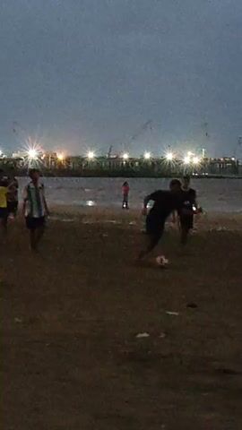 A group of young men are playing soccer on a beach at dusk. The sky is overcast and the lighting is dim, with artificial lights visible in the distance along a pier or shoreline. The men are dressed in casual clothing, some wearing t-shirts and shorts. One man in the foreground is dribbling a soccer ball, while others are positioned to receive passes or defend. The beach appears to be sandy with some debris scattered on the ground. The ocean is visible behind the players, with small waves lapping at the shore.