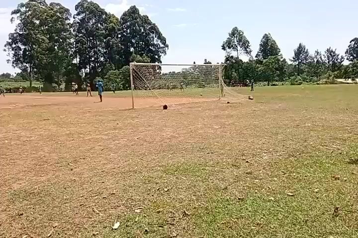 A wide outdoor shot of a soccer field with two goalposts. On the field, there are several players dressed in blue and green uniforms. The field is mostly dry with patches of green grass. In the background, there are numerous trees under a bright sky. The video shows the soccer field and players from a stationary camera position.