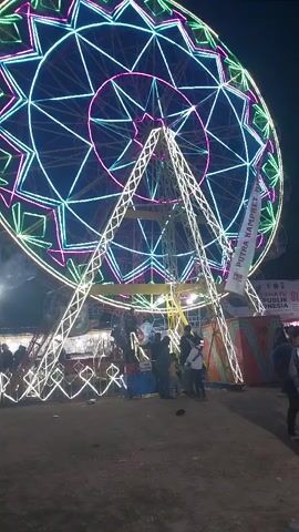 This is a nighttime video of an outdoor fair or carnival. In the foreground and center, a large Ferris wheel is lit with colorful neon lights, primarily white, green, and pink, forming a starburst pattern. The wheel has a sturdy metal support structure also adorned with white lights. Several people are visible standing around the base of the Ferris wheel and in the background, appearing to be patrons of the fair. Some are walking, and others are stationary. Further in the background, other carnival rides and stalls are dimly lit and blurred due to motion. A flag with text is attached to the Ferris wheel's support structure. The ground appears to be dirt or gravel. The overall atmosphere is festive and vibrant due to the bright lights of the attractions.