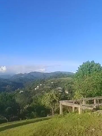 The video shows a scenic view of rolling green hills and mountains under a bright blue sky with scattered white clouds. The foreground consists of a grassy area with lush green trees. In the midground, densely forested hills stretch into the distance, with occasional small houses or buildings visible among the trees. Further in the background, the tops of distant mountains can be seen, some of which are capped with snow. The camera slowly pans from right to left, revealing more of the mountain range and the vastness of the landscape.