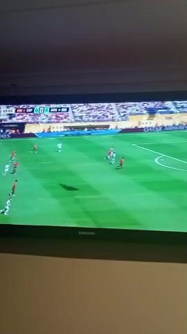 The video shows a screen displaying a soccer game. The score is 0-0 between ESP and ARG. The game is in its 3rd minute. Players in white uniforms are on the field, with some in red uniforms. The field is green, and there are spectators in the background. The screen is mounted on a wall.