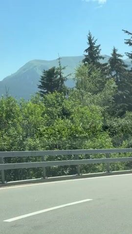 The video shows a view from a moving vehicle on a paved road. A metal guardrail runs along the right side of the road. Beyond the guardrail, there is lush green vegetation, including numerous tall evergreen trees. In the background, majestic mountains with snow-capped peaks rise against a bright blue sky dotted with white, fluffy clouds. The road appears to be in a mountainous, forested region during the daytime.