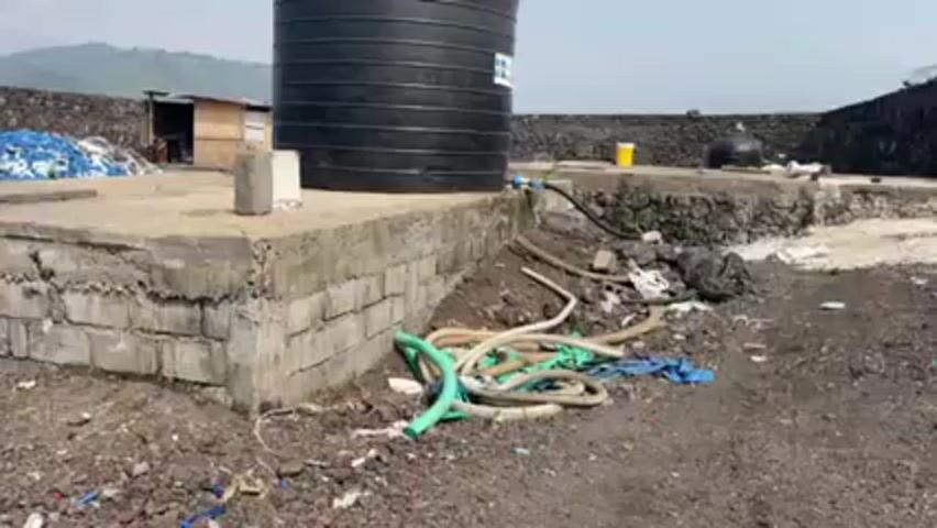 A stationary shot captures a scene of environmental concern. At the center stands a large black water tank, supported by a concrete platform constructed with cinder blocks. A cluster of tangled green and beige hoses lies on the ground at the base of the platform, along with scattered pieces of blue cloth and other debris. In the background, a structure made of black rocks is visible, with a wire fence running along the top. Various containers and plastic items are scattered around this structure. To the left, a small building with a sloped roof is present, partially obscured by a pile of blue material. The ground is dark and appears to be a mix of soil and rocks, further littered with bits of plastic and other trash. The overall impression is one of neglect and environmental degradation.