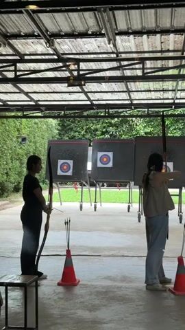 The video shows two individuals practicing archery under a covered structure with a metal roof. On the left, a person in a black shirt and dark pants stands on a low platform, holding a recurve bow in their left hand and drawing it back with their right. An arrow is nocked on the string. In the center foreground, an orange traffic cone holds several arrows. To the right, a second person in a tan sleeveless top and light blue jeans stands facing away from the camera, also holding a recurve bow and drawing it back with an arrow nocked. In the background, three large, square archery targets are mounted on stands. The environment appears to be an outdoor archery range with green foliage visible beyond the targets.