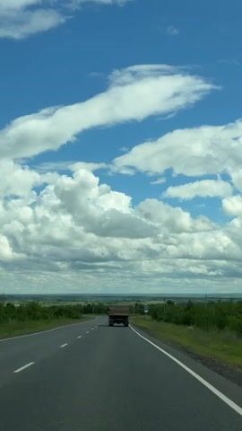 The video shows a rear view from a vehicle traveling on an asphalt road. The road has white dashed lines marking the lanes. On the right side of the road, there is a green grassy shoulder with some vegetation. In the distance, the landscape appears to be flat and covered with trees and fields. A large truck is driving in front of the vehicle. The sky is blue with white, fluffy clouds. There are some road signs visible on the right side of the road. The video appears to be recorded during the daytime.