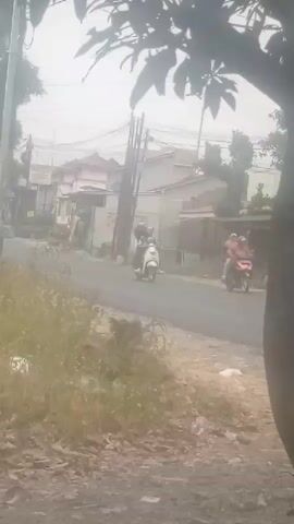 The video shows a residential street with houses and trees. A scooter carrying two people drives past on the road. A dark-colored pickup truck with a tarp covering its bed drives by. The sky is overcast.
