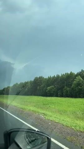 The video shows a view from inside a car driving down a paved road. On the right side of the road, there is a wide, green grassy field bordered by a dense line of tall, green trees. The sky is overcast with dark, heavy clouds, suggesting an impending storm. A road sign with a white arrow indicating a diverging path is visible on the right side of the road at the 00:02 mark. The left side of the car's side mirror is visible in the lower left corner of the frame.